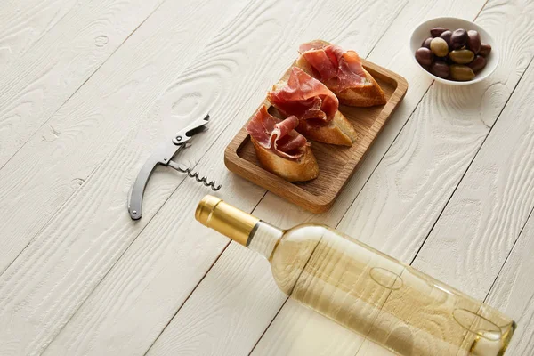 Bottle with white wine near prosciutto on baguette, olives and corkscrew on white wooden surface — Stock Photo