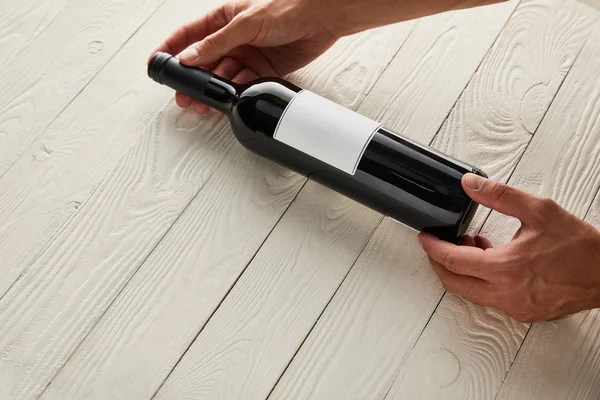 Cropped view of man holding bottle of wine with blank label on white wooden surface — Stock Photo