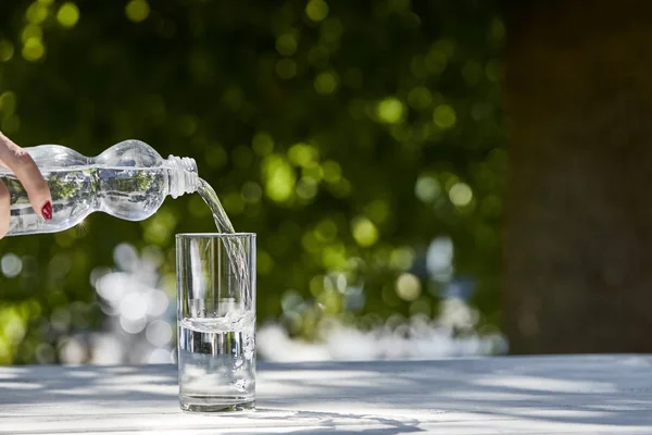 Vue recadrée de la femme versant de l'eau fraîche et propre de la bouteille en verre transparent à la journée ensoleillée sur une table en bois — Photo de stock
