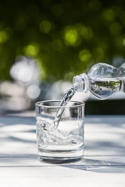 Eau propre fraîche coulant de la bouteille en verre transparent à la journée ensoleillée sur une table en bois — Photo de stock