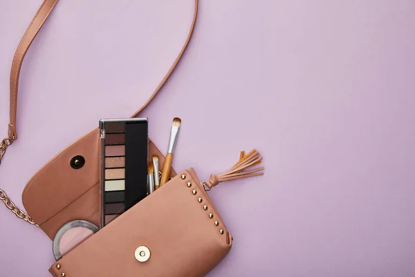 Top view of eye shadow and blush near cosmetic brushes in bag isolated on violet — Stock Photo