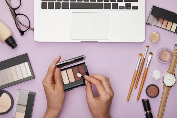 Vue recadrée de la femme tenant ombre à paupières près d'un ordinateur portable et des cosmétiques décoratifs isolés sur violet — Photo de stock