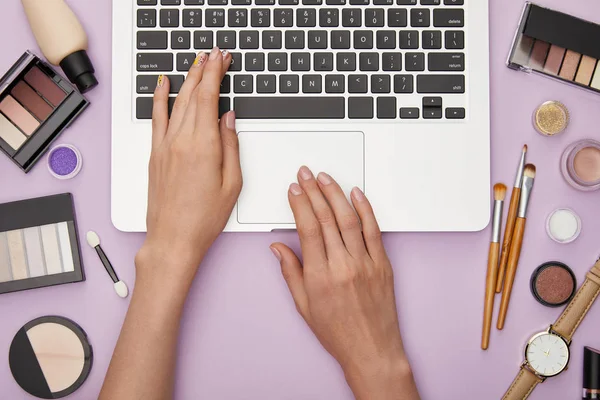 Vista superior de la mujer escribiendo en el ordenador portátil cerca de cosméticos decorativos aislados en violeta - foto de stock