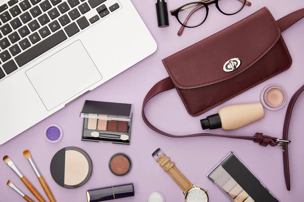 Draufsicht auf Tasche und dekorative Kosmetik in der Nähe von Laptop isoliert auf violett — Stockfoto