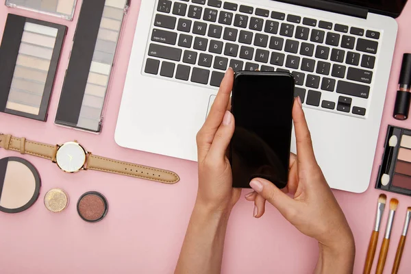 Vista superior de la mujer que sostiene el teléfono inteligente con la pantalla en blanco cerca del ordenador portátil y cosméticos decorativos aislados en rosa — Stock Photo
