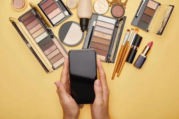 Vista recortada de la mujer sosteniendo teléfono inteligente con pantalla en blanco cerca de cosméticos decorativos aislados en amarillo - foto de stock