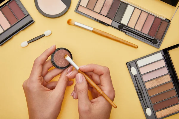 Top view of woman holding cosmetic brush near eye shadow isolated on yellow — Stock Photo