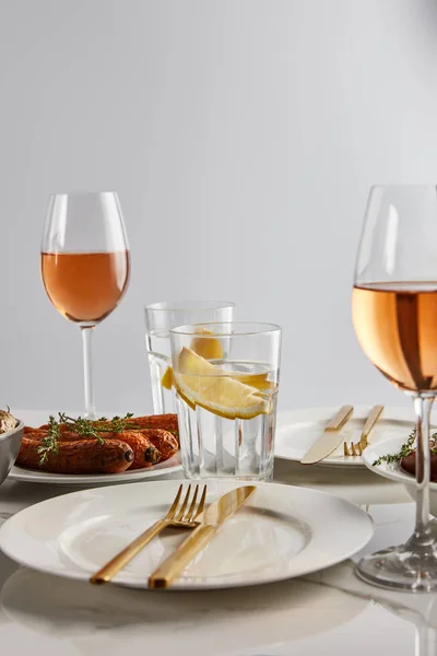 Zanahorias al horno, vasos con vino de rosas y agua de limón, platos blancos y cubiertos de oro en la mesa de mármol aislado en gris - foto de stock