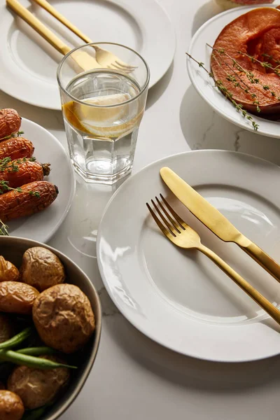Platos blancos con verduras horneadas, tenedores dorados y cuchillos, vaso con agua de limón sobre mesa de mármol blanco - foto de stock