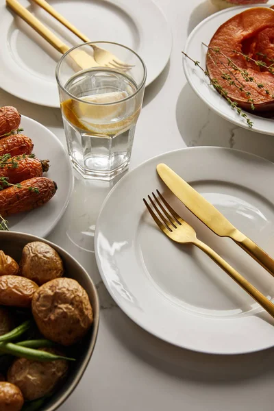 White plates with baked vegetables, golden forks and knives, glass with lemon water on white marble table — Stock Photo