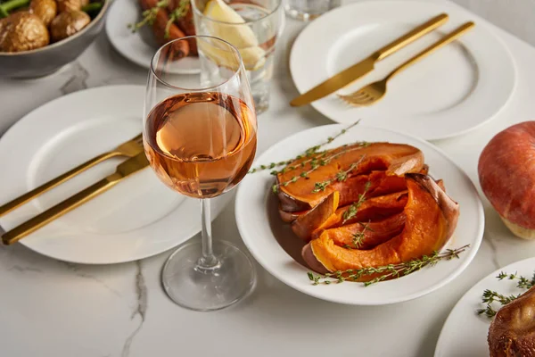 Vaso con vino rosa cerca de calabaza cortada al horno y platos con tenedores y cuchillos sobre mesa de mármol - foto de stock