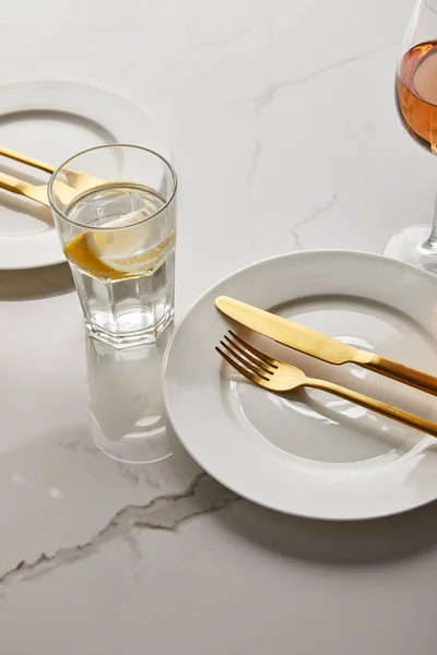 Стекло с лимонной водой возле белых тарелок с золотыми вилами и ножами на мраморном столе — стоковое фото
