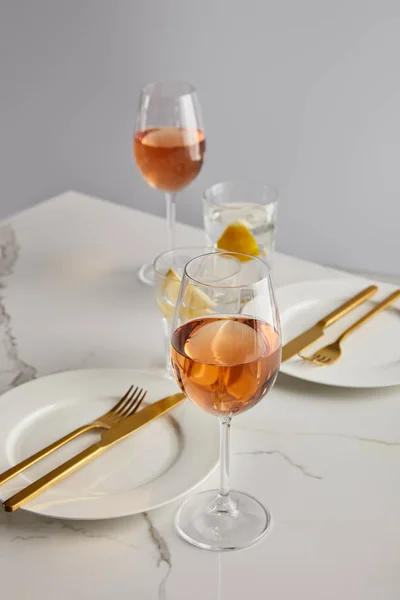 Vasos con vino de rosas y agua de limón cerca de platos blancos con tenedores y cuchillos sobre mesa de mármol aislados en gris - foto de stock
