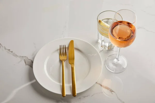 Plato blanco con cuchillo de oro y tenedor cerca de vasos con agua de limón y vino rosa sobre mesa de mármol - foto de stock