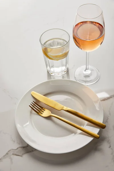 Plato blanco con cuchillo de oro y tenedor cerca de vasos con agua de limón y vino rosa sobre mesa de mármol - foto de stock