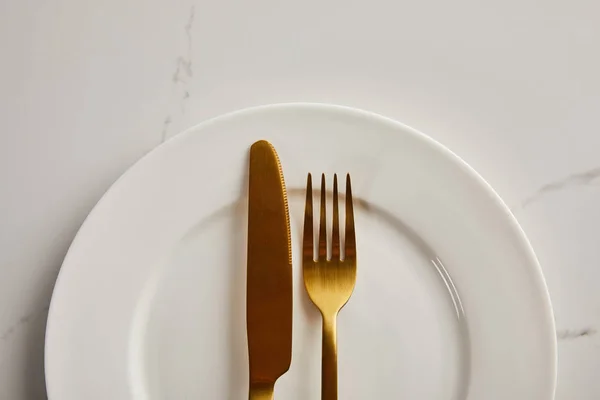 Vue du dessus du couteau et de la fourchette dorés sur une plaque blanche propre sur une table en marbre — Photo de stock