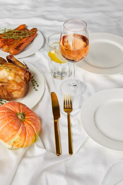 Целая тыква, жареная цыпленок и запечённая морковь возле стаканов с розовым вином и лимонной водой на белой скатерти — стоковое фото