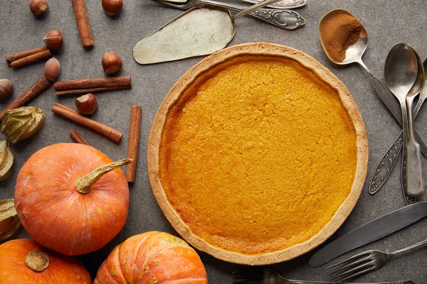 Top view of delicious pumpkin pie near whole pumpkins, cutlery, hazelnuts and cinnamon sticks on grey stone surface — Stock Photo