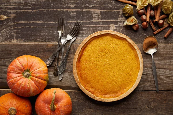 Vue de dessus de délicieuse tarte à la citrouille près de citrouilles fraîches entières et couverts de surface en bois sombre — Photo de stock