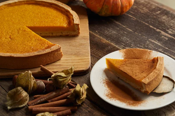 Delicious pumpkin pie with cinnamon powder near physalis and cinnamon sticks on dark wooden surface — Stock Photo