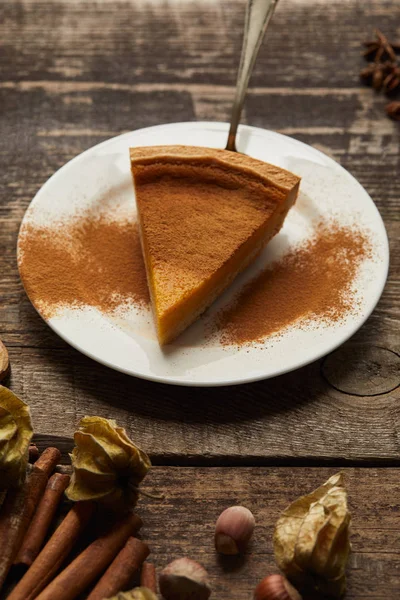 Delicious pumpkin pie with cinnamon powder on white plate near cinnamon sticks and physalis on dark wooden table — Stock Photo