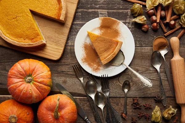 Vue de dessus de tarte à la citrouille savoureuse sur assiette avec spatule près de citrouilles entières et couverts sur une surface en bois sombre — Photo de stock