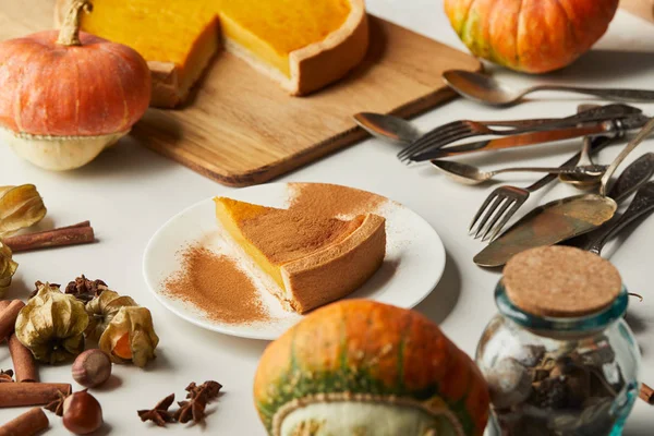 Sabroso pastel de calabaza cerca de calabazas enteras, cubiertos y especias en la superficie blanca - foto de stock