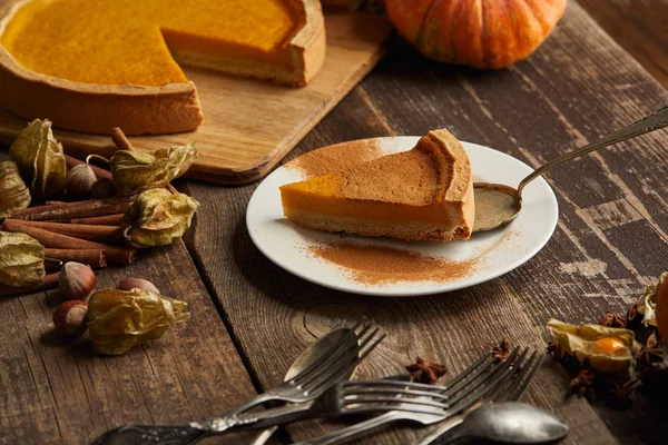 Delicious pumpkin pie with cinnamon powder near cutlery and spices on dark wooden surface — Stock Photo