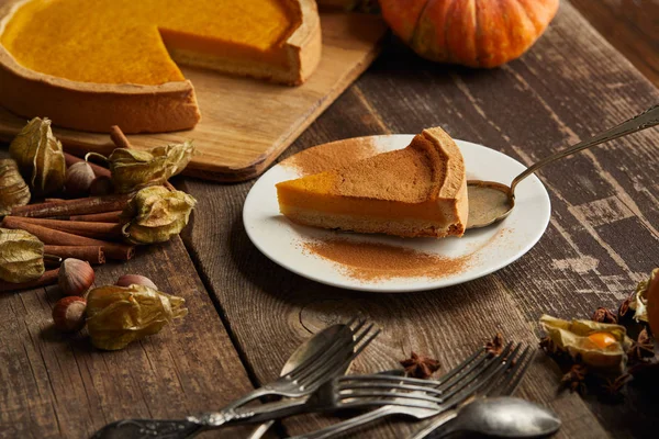 Délicieuse tarte à la citrouille avec de la poudre de cannelle près des couverts et des épices sur une surface en bois sombre — Photo de stock