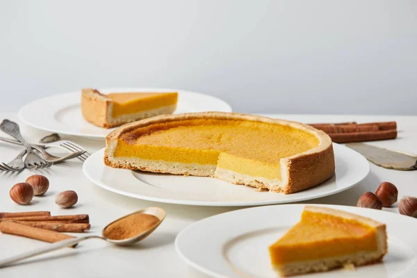 Белые тарелки с вкусным тыквенным пирогом рядом со столовыми приборами и специями, изолированными на сером — стоковое фото