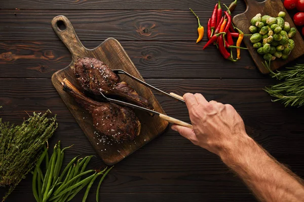 Vue recadrée de l'homme prenant de la viande savoureuse de la planche à découper avec une pince à épiler — Photo de stock