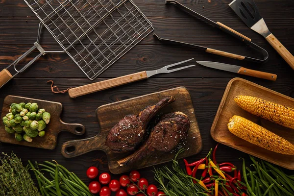 Blick von oben auf Fleisch, Mais, Chilischoten, Kirschtomaten, grüne Erbsen, Grün, Rosenkohl auf Schneidebrett und Grillwerkzeug — Stockfoto