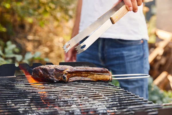 Vista recortada del hombre con pinzas parrilla carne en la parrilla de barbacoa - foto de stock