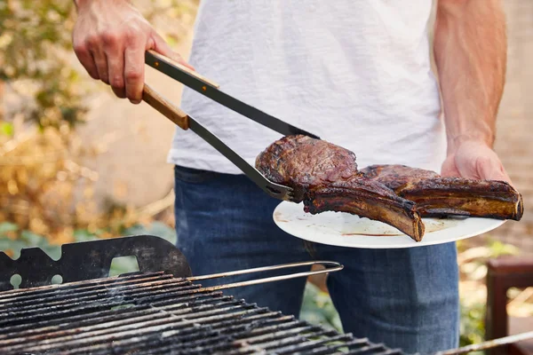 Vista recortada del hombre con pinzas que sostienen la carne en el plato - foto de stock