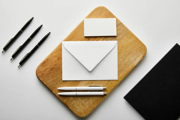 Vista superior de sobre, tarjeta de visita y bolígrafos blancos en tablero de madera, cuaderno y bolígrafos negros en la mesa - foto de stock