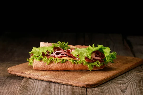 Sándwich fresco con lechuga, jamón, queso, tocino y tomate sobre tabla de cortar de madera aislada en negro - foto de stock