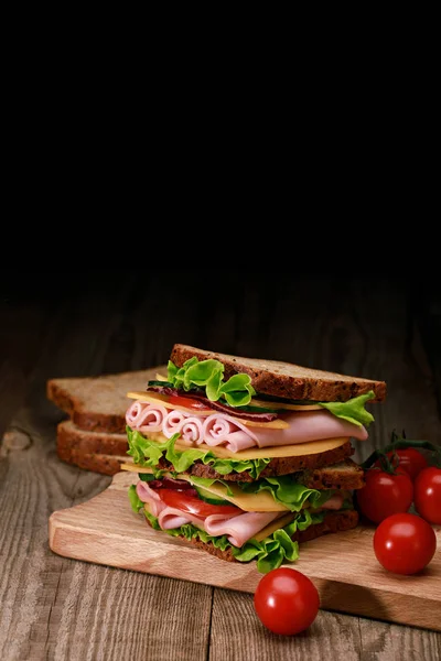 Fresh sandwich with lettuce, ham, cheese, bacon and tomato on wooden cutting board with cherry tomatoes isolated on black — Stock Photo