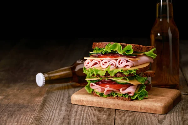 Fresh sandwich with lettuce, ham, cheese, bacon and tomato on wooden cutting board near bottles of beer isolated on black — Stock Photo