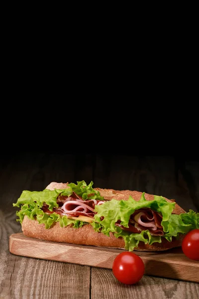 Fresh sandwich with lettuce, ham, cheese, bacon on wooden cutting board with cherry tomatoes isolated on black — Stock Photo
