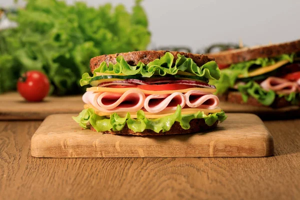 Sándwich fresco con lechuga, jamón, queso, tocino y tomate sobre tabla de cortar de madera aislada en gris - foto de stock