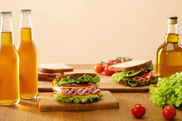 Foco seletivo de sanduíches frescos com alface, presunto, queijo, bacon e tomate perto de garrafas de cerveja em mesa de madeira isolada em bege — Fotografia de Stock