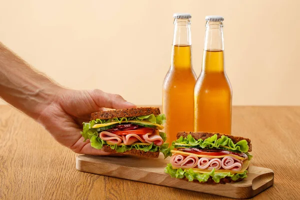 Обрезанный вид человека, держащего свежий сэндвич с салатом, ветчиной, сыром, беконом и помидорами рядом с бутылками пива за деревянным столом, изолированным на бежевом — стоковое фото