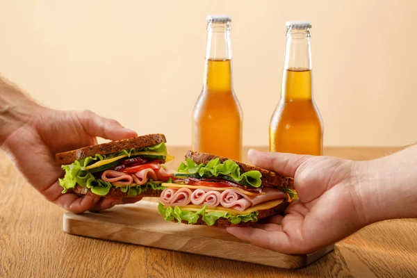 Vista recortada de hombres sosteniendo sándwiches frescos con lechuga, jamón, queso, tocino y tomate cerca de botellas de cerveza en la mesa de madera aislada en beige - foto de stock