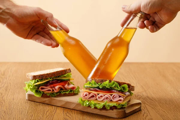 Vista recortada de hombres sosteniendo botellas de cerveza cerca de sándwiches frescos con lechuga, jamón, queso, tocino y tomate cerca en mesa de madera aislada en beige - foto de stock