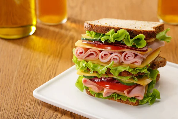 Focus selettivo di panino fresco con lattuga, prosciutto, formaggio, pancetta e pomodoro vicino a bottiglie di birra al tavolo di legno — Foto stock