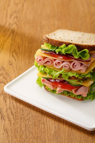 Свежий сэндвич с салатом, ветчиной, сыром, беконом и помидорами за деревянным столом — стоковое фото