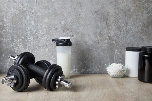 Shake de protéine dans la bouteille de sport près des haltères et poudre de protéine près de mur de béton — Photo de stock