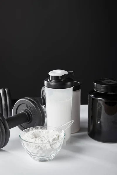 Glass bowl near dumbbells and sports bottle with protein shake on black — Stock Photo