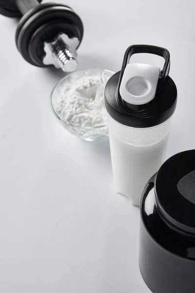 Селективный фокус спортивной бутылки с протеиновым коктейлем возле банки и гантели на белом — стоковое фото