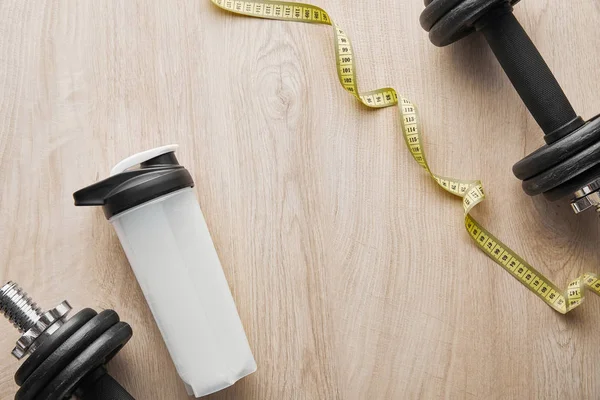 Top view of dumbbells near sports bottle with protein shake near measuring tape on wooden surface — Stock Photo